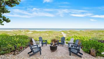 A patio with Adirondack chairs, a fire pit, and a view of the beach and dunes.