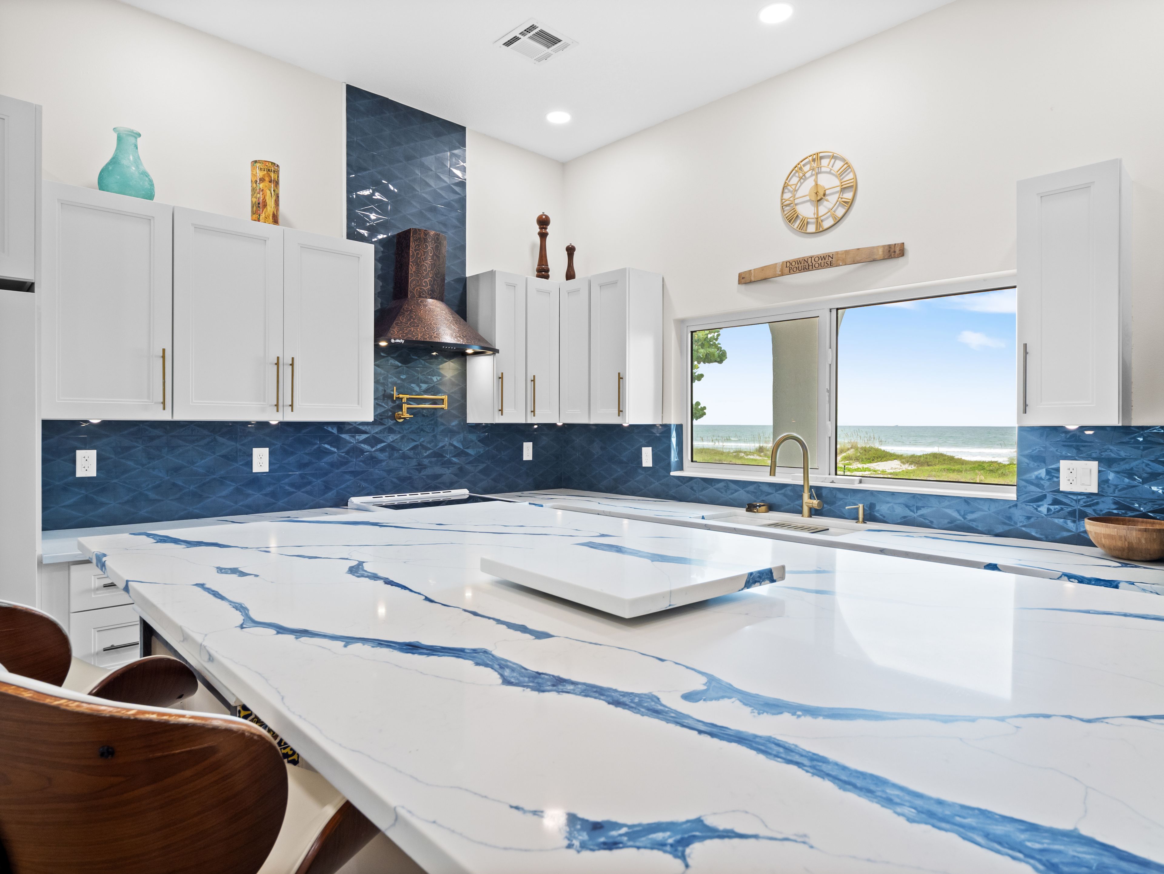 A modern kitchen with a large island, white cabinets, blue backsplash, and a view of the ocean.