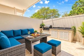 An outdoor patio with a dining table, blue cushioned seating, a grill, and a view of trees.