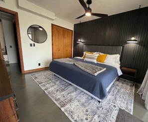 A bedroom with a bed, a ceiling fan, a round mirror, and a wooden door.
