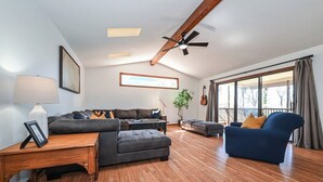 A spacious living room with a wooden floor, a ceiling fan, a large window with curtains, a dark-colored sofa, a blue armchair, and a framed picture on a side table.
