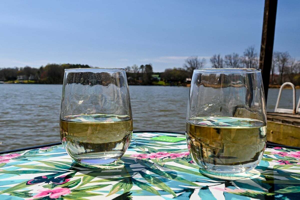 Two glasses of white wine on a table with a floral pattern, set against a backdrop of a lake and trees.