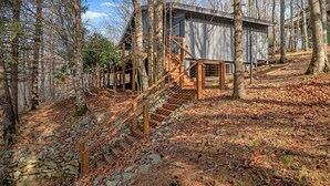 A rustic cabin with a wooden staircase leading to a stone-walled entrance, surrounded by trees and a leaf-covered ground.