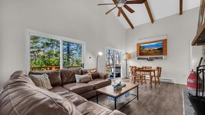 A modern living room with a brown leather sofa, a coffee table, and a dining area with wooden chairs. There is a ceiling fan, a TV mounted on the wall, and a view of trees through large windows.