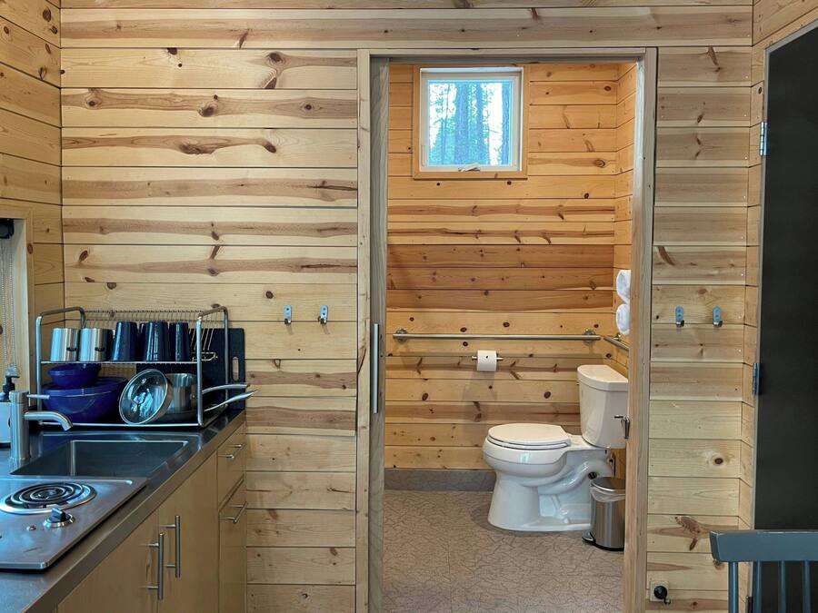L’intérieur d’une cabane en bois comprend un coin cuisine équipé d’une cuisinière, d’un évier et d’un porte-ustensiles. On aperçoit les toilettes et une fenêtre dans la salle de bain adjacente.