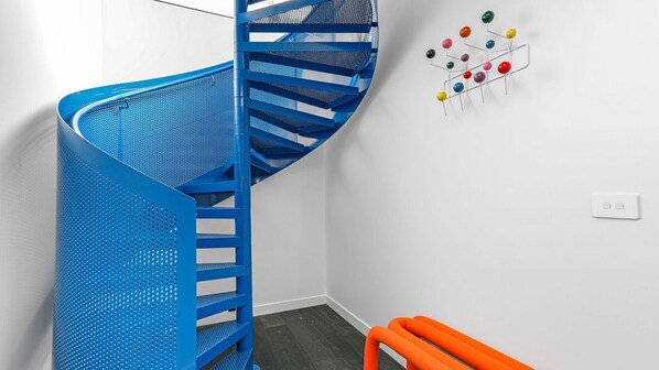 A blue spiral staircase in a room with white walls, a small orange bench, and a wall-mounted color wheel.