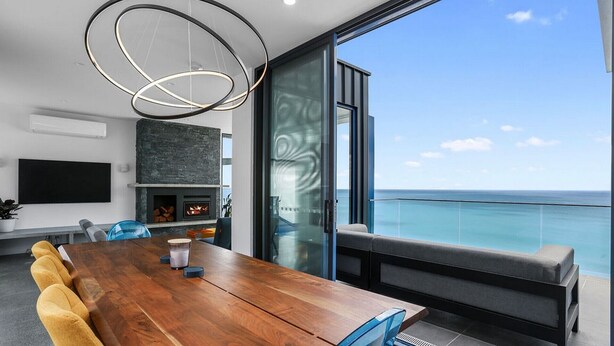 Una sala de estar moderna con una mesa de comedor de madera, una chimenea y un gran ventanal que da a un balcón con vista al mar.