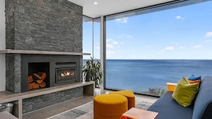 Una sala de estar moderna con chimenea de piedra, un ventanal con vistas al mar y un sofá azul con cojines de colores.