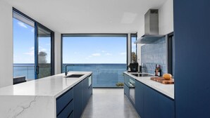 Una cocina moderna con un ventanal grande con vistas al mar, horno empotrado y encimera con tabla de cortar y botellas.