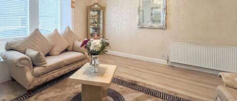 A living room with a beige sofa, a coffee table, a patterned rug, a chandelier, a mirror, and a radiator.