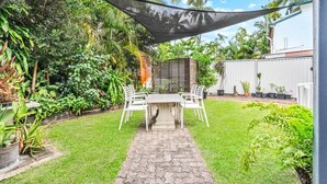 Une terrasse avec une table et des chaises, entourée d’une végétation luxuriante et d’une clôture blanche.