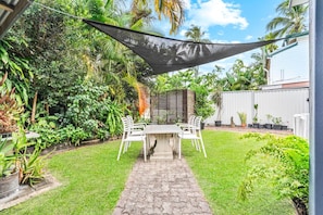 A patio with a table and chairs, surrounded by lush greenery and a white fence.
