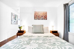 Une chambre avec un grand lit, deux tables de chevet, une œuvre d’art murale et une fenêtre avec des rideaux.