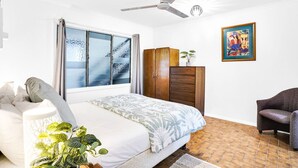 A bedroom with a bed, a wardrobe, a chair, a ceiling fan, a window with curtains, and a painting on the wall.