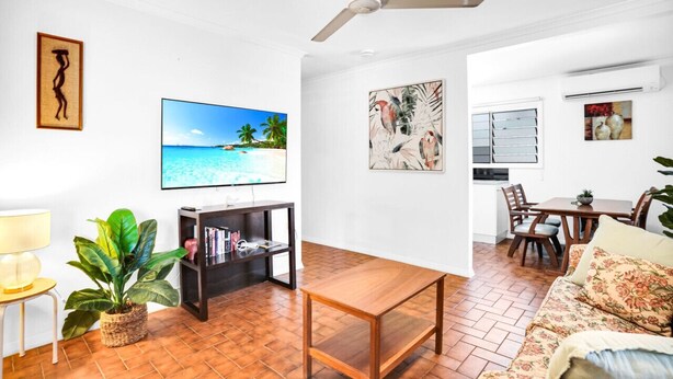 A living room with a TV displaying a beach scene, a ceiling fan, a dining area with a table and chairs, and a kitchen area with a window and a plant.
