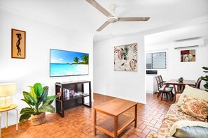 A living room with a TV displaying a beach scene, a ceiling fan, a dining area with a table and chairs, and a kitchen area with a window and a plant.