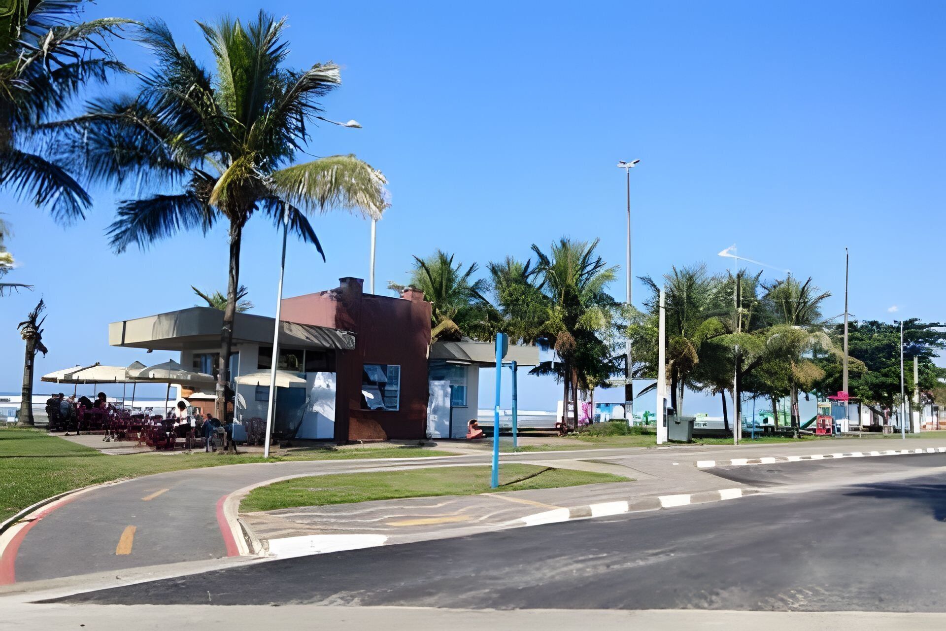 A coastal road with a building that has a red roof and a blue structure. Palm trees line the street, and there is a clear blue sky.