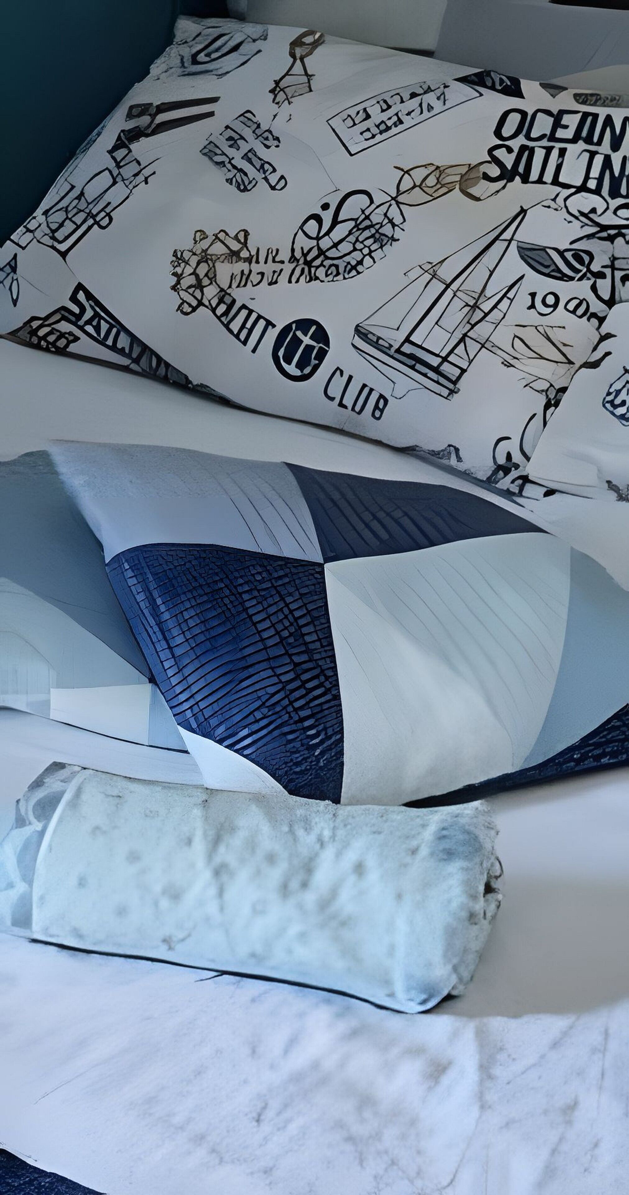 A bed with a white pillow featuring nautical illustrations and text, a blue and white patterned pillow, and a rolled-up white blanket.