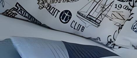 A bed with a white pillow featuring nautical illustrations and text, a blue and white patterned pillow, and a rolled-up white blanket.