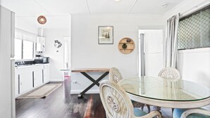 Una cocina moderna con armarios blancos, microondas y suelo de madera. Cuenta con una zona de comedor con mesa de vidrio y sillas de mimbre.