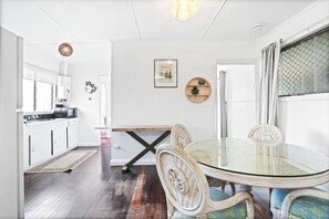 A modern kitchen with white cabinets, a microwave, and a wooden floor. There is a dining area with a glass table and wicker chairs.
