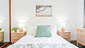 Un dormitorio ordenado con cama, dos mesitas de noche, una mesita auxiliar, un cuadro en la pared y una ventana con cortinas.