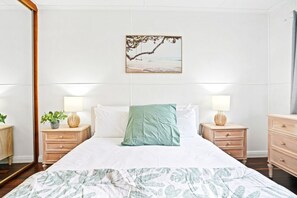 A neatly arranged bedroom with a bed, two bedside tables, a nightstand, a wall-mounted picture, and a window with curtains.