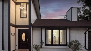 Une maison à deux étages avec une porte marron foncé, un lustre allumé et une fenêtre équipée de stores.