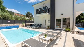 A modern pool area with lounge chairs and a small table, a stone wall, and a clear blue sky.