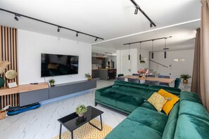 A modern living room with a large flat-screen TV, a green sofa, a coffee table, and a kitchen area in the background. - Villa Marevia by Aneo Travel (Baska)