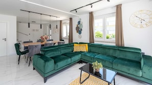 A modern living room with a green sectional sofa, a dining area with a wooden table, and a large wall clock.