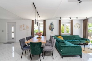 A modern living room with a dining area, green sofa, and a large window with curtains. - Villa Marevia by Aneo Travel (Baska)