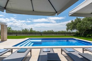En rektangulär swimmingpool med ett klart, blått vatten, omgiven av en kakelbeklädd terrass. Här finns solstolar med handdukar och ett stort parasoll som ger skugga.