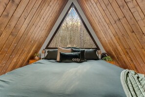 A triangular wooden room with a bed, pillows, a small table, and a window. - Evergreen Dreams | A-Frame Retreat + Hot Tub/Sauna (Polk County)