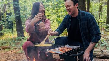 Una pareja cocina al aire libre junto a una fogata, rodeada de un bosque.
