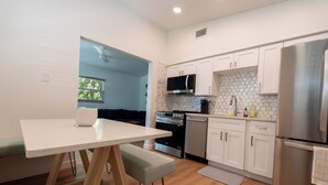 Una cocina moderna con armarios blancos, un refrigerador de acero inoxidable, un microondas y un horno. Hay una mesa de comedor con un banco, y una ventana con vista a árboles.
