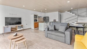 Ett modernt vardagsrum med en grå soffa, en gul fåtölj, en platt-TV och en köksdel med mikrovågsugn och ugn.
