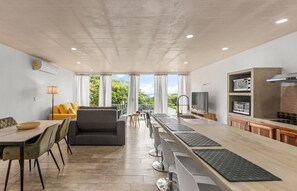 Et moderne køkken med en lang bordplade, et spiseområde med en gul sofa, og udsigt til grøn vegetation udenfor.