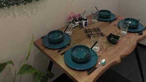Mesa de comedor dispuesta con platos verde azulado, vasos y cubiertos, rodeada por un muro de follaje verde y un letrero de neón.