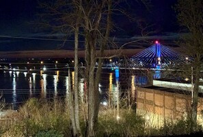 Une vue nocturne d’un pont dont les câbles sont illuminés, avec une étendue d’eau au premier plan et des arbres.