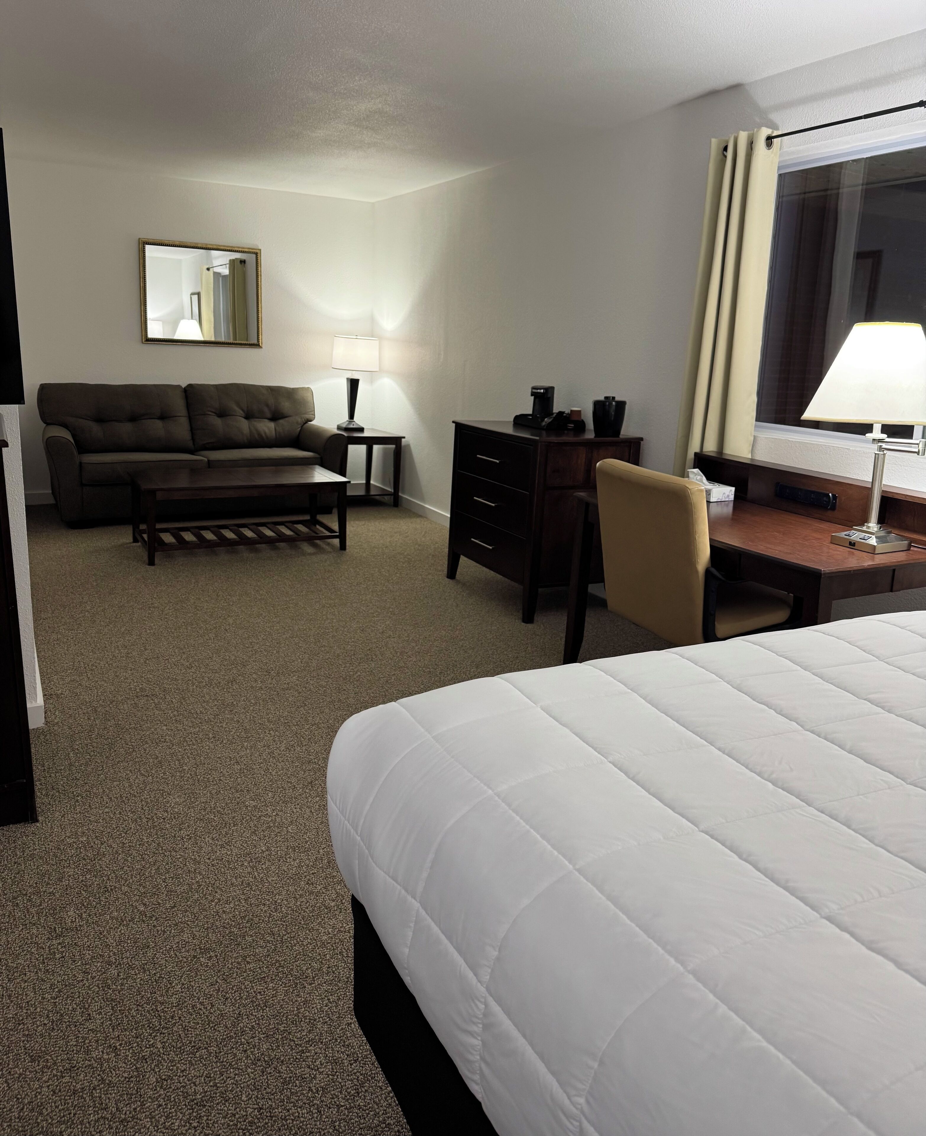 A hotel room with a large bed, a sofa, a desk, a chair, a coffee maker, and a lamp.