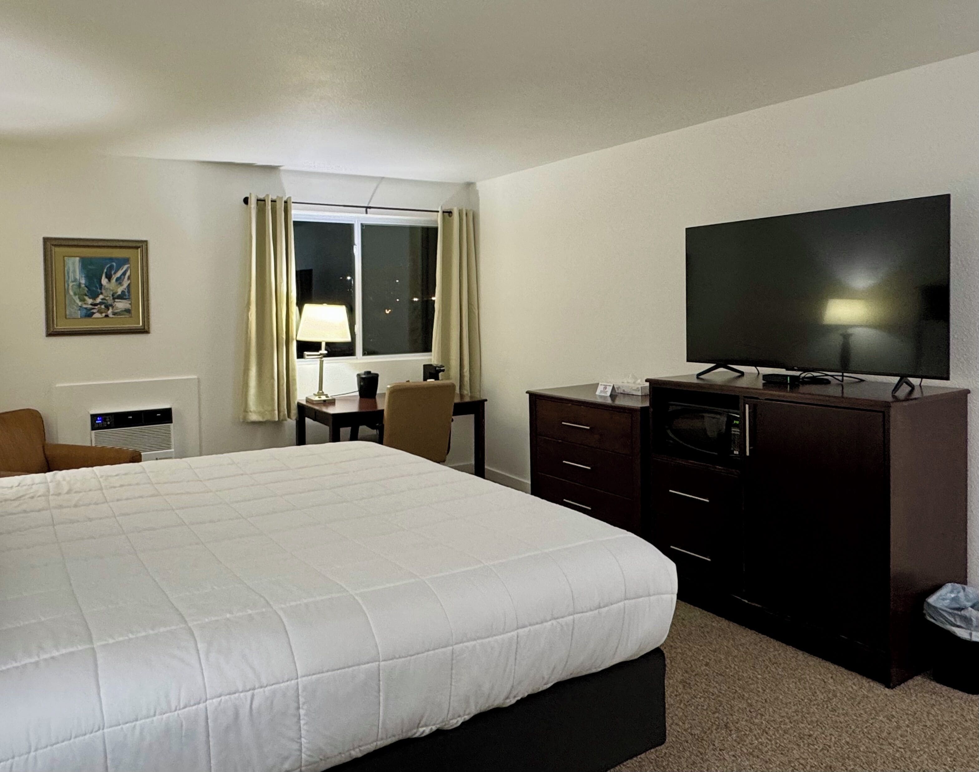 Quarto de hotel com uma cama grande, televisão de tela plana, escrivaninha com luminária e janela com cortinas.