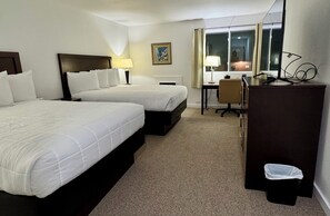 Une chambre d’hôtel avec deux lits, un bureau, une chaise, une poubelle, une fenêtre et un tableau au mur.