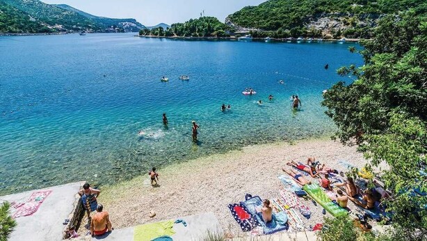 Une plage avec une eau cristalline, des gens qui se baignent et prennent des bains de soleil, et des bateaux à l’horizon.