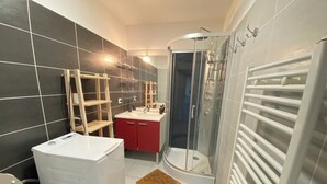 Une salle de bain moderne avec un lave-linge blanc, un espace douche, un meuble-lavabo rouge et un sol carrelé.