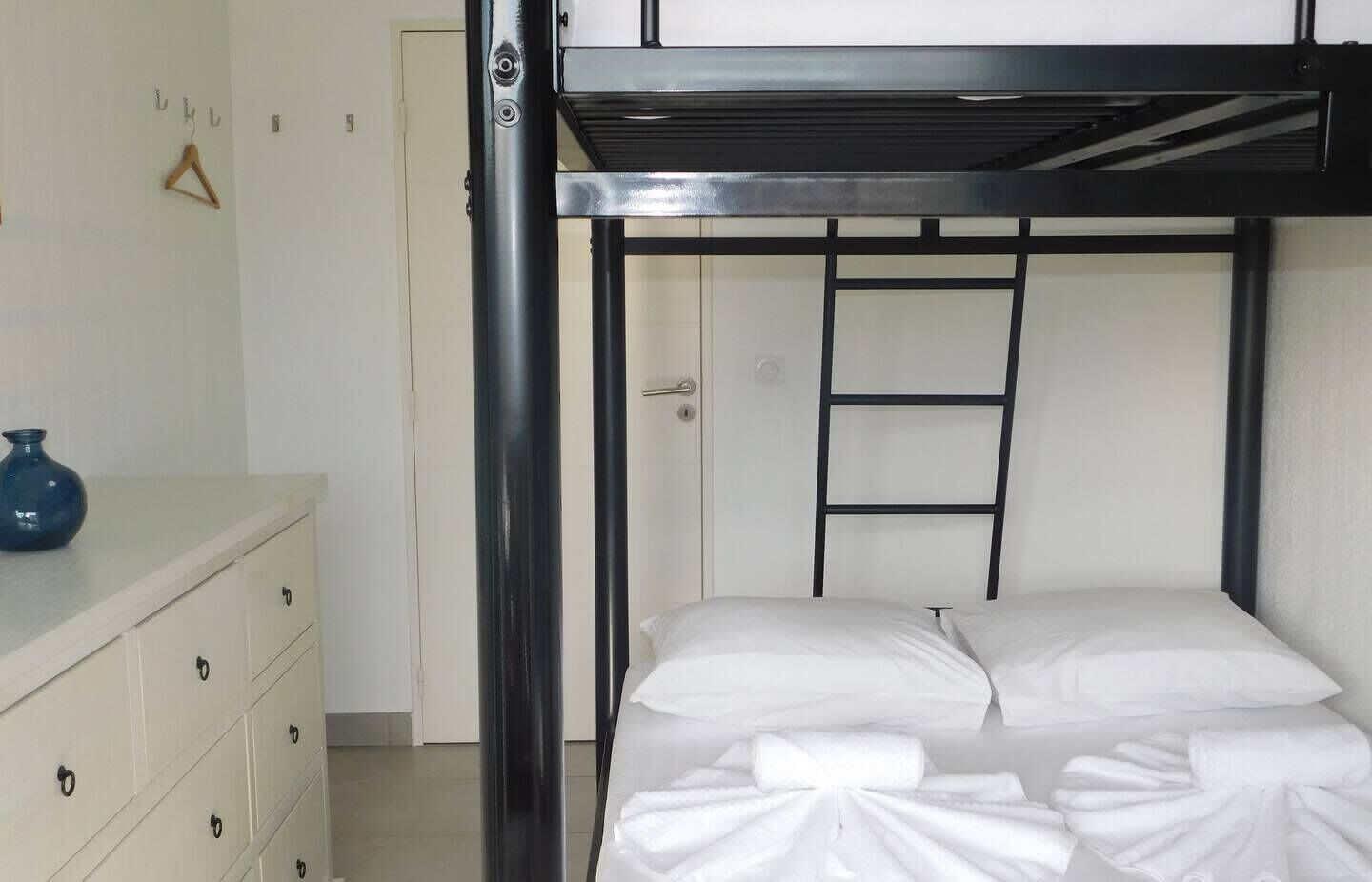 Un lit superposé avec une échelle, une commode avec des tiroirs et un vase bleu posé sur la commode.