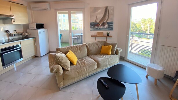 Un appartement moderne doté d’une kitchenette, d’un canapé avec des coussins jaunes, d’une table basse noire et d’un tableau représentant un voilier.