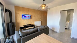 A modern living room with a wooden wall, a black wood-burning stove, a grey sofa, a wooden coffee table, and a TV mounted on the wall.
