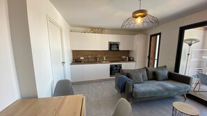 A modern kitchen with white cabinets, a wooden countertop, and a built-in oven. There is a grey sofa and a round coffee table in the living area.