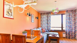 A cozy living room with a wooden dining table, a sofa, and a window with patterned curtains.